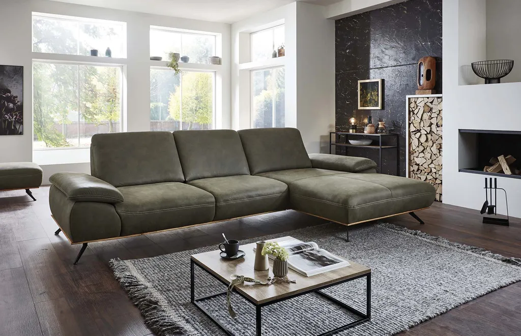 Großes Ecksofa in Olivgrün mit Lederoptik, schwarzen Metallfüßen und gemütlichen Armlehnen. Modernes Wohnzimmer mit Kamin, Holzdeko und großem Teppich – perfekte Kombination aus Komfort und Design.